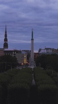 Aerial view gliding above tree lined alley toward Freedom Monument and Saint Peter Church tower in Riga, Latvia during evening twilight.