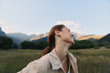 Fototapeta premium Happy woman outdoors in nature, smiling with eyes closed, enjoying fresh air, casual clothing, green meadow and mountains background, peaceful moment