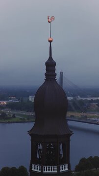 Aerial view of the historic Riga Cathedral spire with its weathercock above the Daugava River and cityscape at dusk in Riga, Latvia.