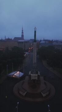 An aerial shot slowly circles the historic Freedom Monument and surrounding park in Riga, Latvia during a dark stormy evening.