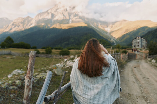 Young woman with long brunette hair walks along a mountain trail, wrapped in a gray shawl as distant peaks loom, a tranquil countryside scene suggesting adventure and contemplation.