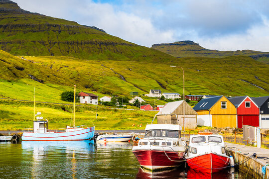 Close-up view of two small motorboats docked alongside a wooden pier in a harbor, framed by green hills under a cloudy sky, Faroe Islands, Denmark