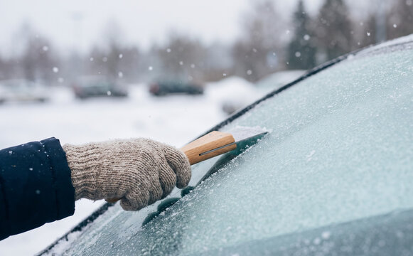 Person in warm mitten scraping ice and snow from a frosted car windshield on a cold winter morning