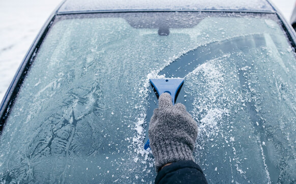 Person scraping frost and ice from a car windshield on a cold winter morning using a plastic tool