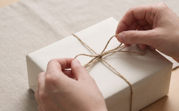 Woman hands tying a bow with twine on a handmade gift box wrapped in white paper for a birthday present