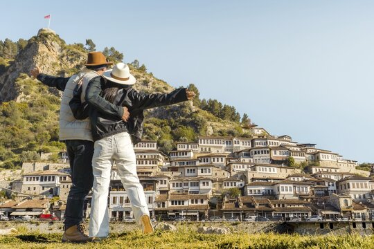 Happy travel couple embracing enjoy arms up vacation visit Mangalem Berat Old Town UNESCO Albania Ottoman architecture hillside houses sunny autumn vacation sightseeing