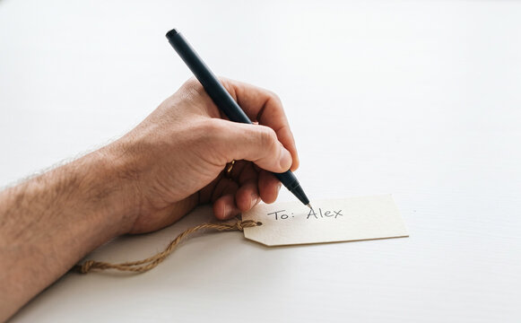 Adult male writing To Alex on a gift tag with a black pen on a white surface