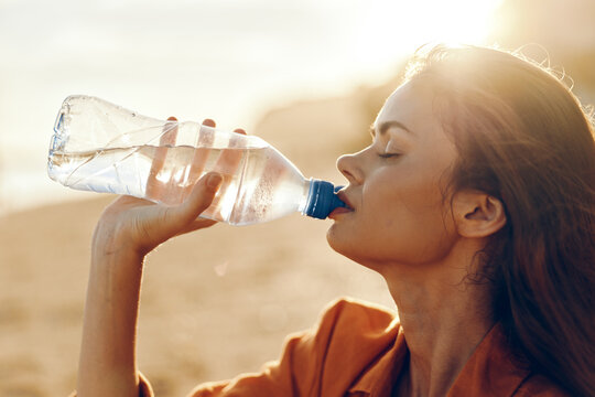 Healthy woman drinking water from plastic bottle outdoors in bright sunlight, warm desert sand background, summer hydration, side profile portrait, wellness and health