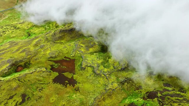 Oblique aerial reveal of moss-covered volcanic plateau as low clouds pull back to expose sinuous shallow stream and peat pools, with moss, stream, plateau, valley detail