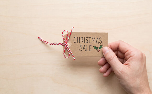 Hand holding a brown paper tag with Christmas sale text and red string on wooden background