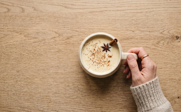A woman holding a warm cup of spicy eggnog with cinnamon and star anise on a wooden table