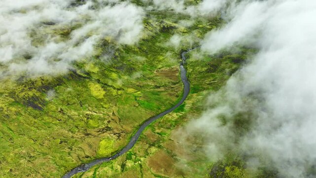 Oblique aerial reveal of moss-covered volcanic plateau as low clouds pull back to expose sinuous shallow stream and peat pools, with moss, stream, plateau, valley detail