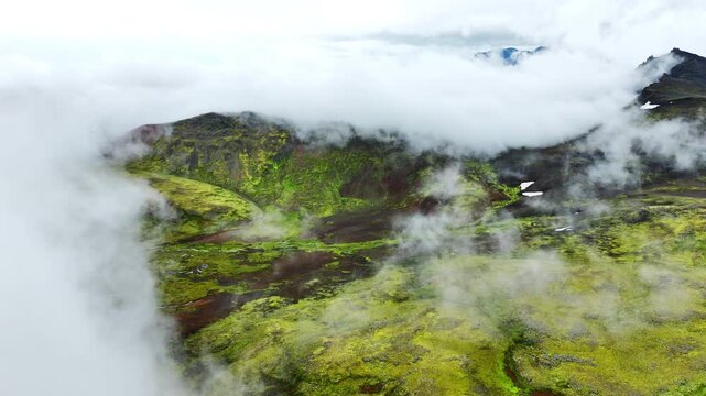 Oblique aerial reveal of moss-covered volcanic plateau as low clouds pull back to expose sinuous shallow stream and peat pools, with moss, stream, plateau, valley detail