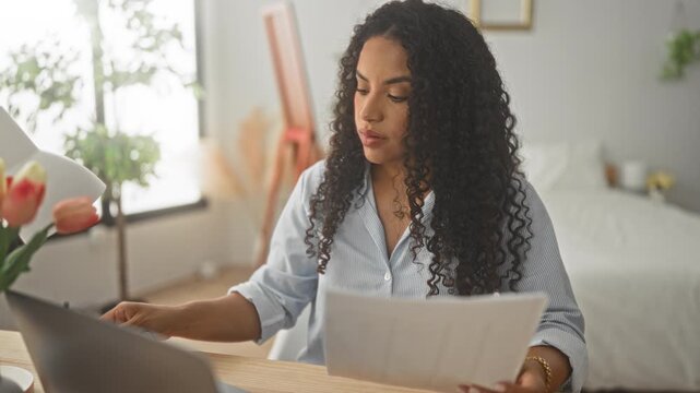 Woman in bedroom adjusting glasses while reading document at laptop on desk; concentration productivity efficiency.