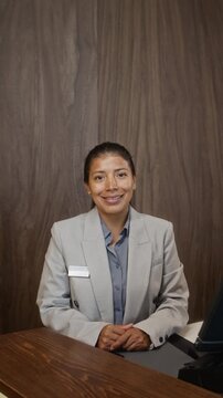 Portrait of Hispanic young adult female receptionist sitting at wooden desk, smiling at camera. Video is ideal for hospitality and customer service marketing campaigns, vertical