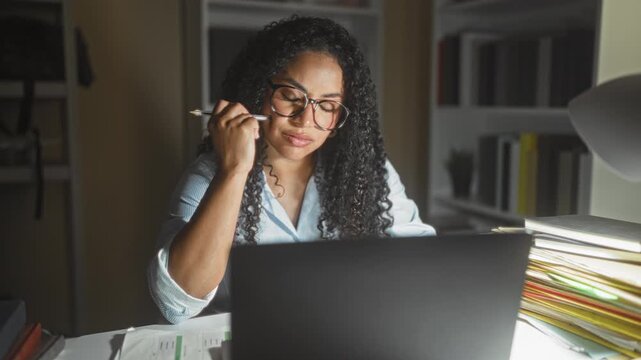 Woman with curly hair and glasses working at laptop, holding pen to temple, surrounded by folders and lamp in office building; focus.
