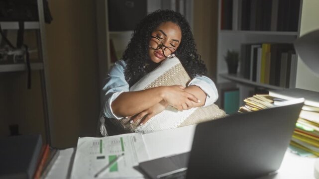 Young woman hugs pillow and adjusts glasses at desk with laptop and paperwork in building; late night fatigue.
