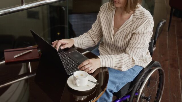 Young adult Caucasian woman with disability sitting in wheelchair and typing on laptop in cafe. High angle shot ideal for corporate diversity, remote work, and inclusive employment campaigns