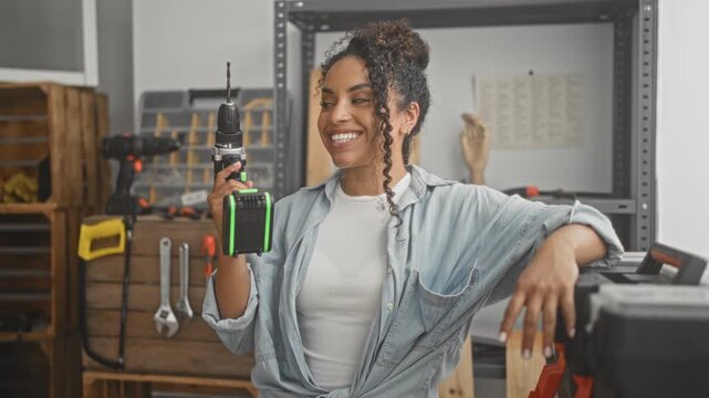 Woman holding a cordless drill upright, right hand gripping the drill and left hand resting on toolbox inside a workshop building; confidence craft skill.