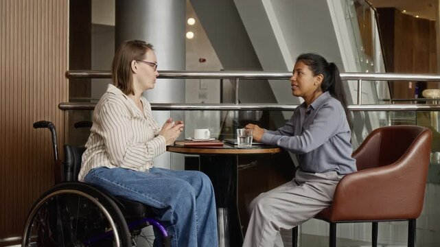 Caucasian young adult woman with disability in wheelchair and Hispanic colleague talking at cafe table. Video promoting workplace diversity and social inclusion for corporate marketing