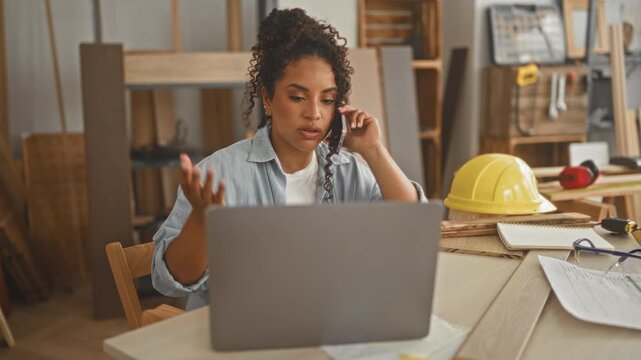 Woman carpenter on phone at studio with laptop and raised hand talking gesture while planning woodworking project; professional focus.