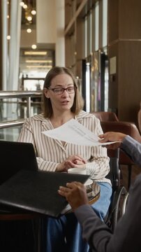 Vertical shot of Caucasian woman with disability in wheelchair reviewing document with Hispanic colleague in cafe. Clip is ideal for inclusive corporate recruitment campaigns