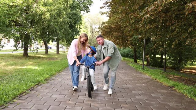 Happy parents teaching their son to ride bicycle outdoors, camera moving backwards. Child safety