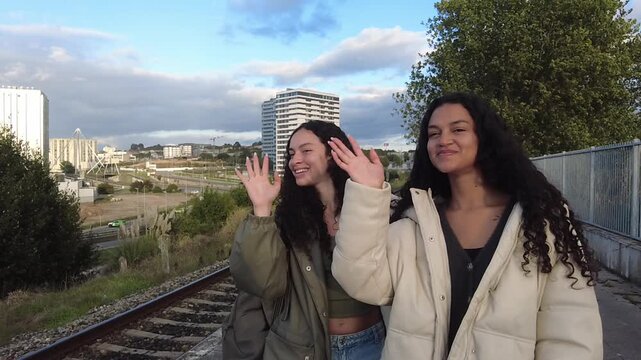 Two beautiful young twin sisters with curly hair smiling and waving goodbye while standing on a train station platform with their luggage, ready for a new journey and adventure together