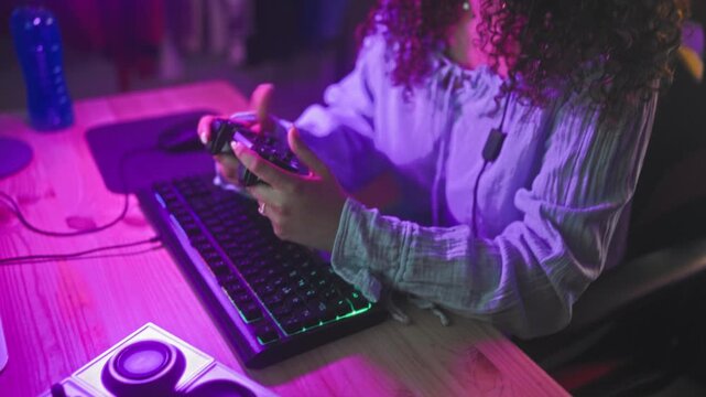 Woman with headset gripping controller playing video games at a computer desk in a studio with neon purple and pink led lights and a gaming chair; concentration.