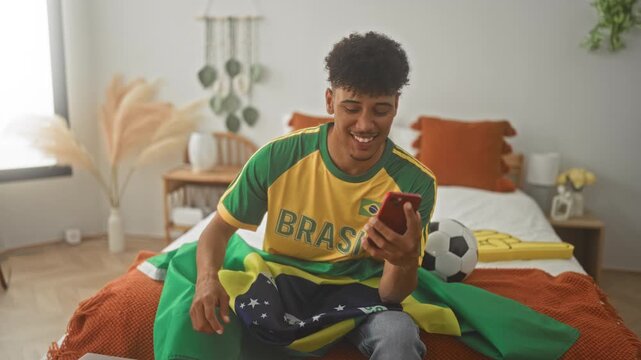 Man in brasil jersey eating pizza slice with hand while holding smartphone on bed inside building; team pride.