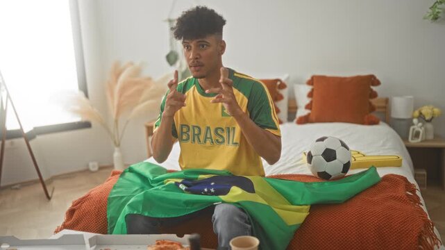 Young man on bed in building wearing yellow brasil jersey with brazil flag and soccer ball, gesturing with hands; national pride excitement.