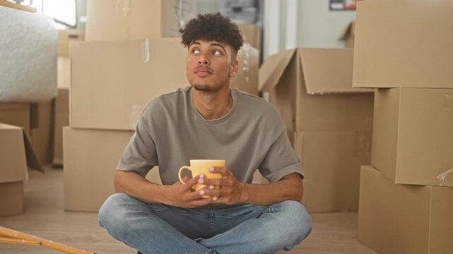 Man holding a yellow mug with both hands, sitting cross legged among packed boxes inside a building; moving day contentment.