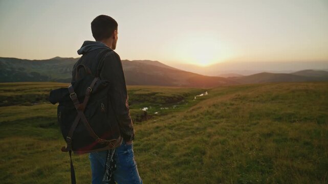 Young man watches the sunset while standing on a grassy hillside near a stream in an open valley