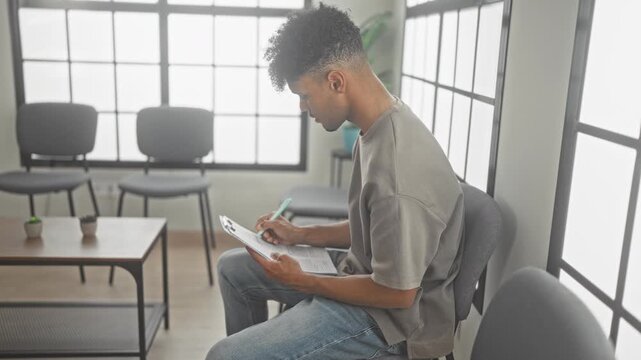 Young hispanic man seated on chair filling out a clipboard form with pen in a clinic waiting room by window and small potted plants; quiet concentration.