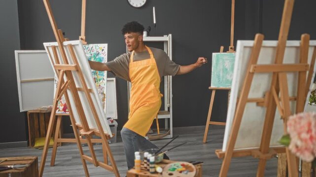 Man painter in yellow apron holds paintbrush in right hand while stepping and dancing beside canvas on studio easel; joyful creativity.