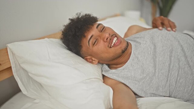 Man lying on bed smiling, reaches arm toward camera with pillow and wooden headboard visible in a bedroom inside a building; morning joy.