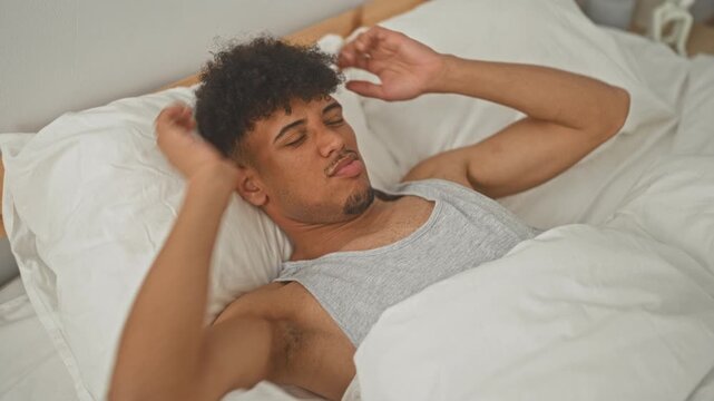 Young hispanic man covers face with hands on bed in bedroom, wearing gray tank top and white duvet visible; fatigue morning headache.