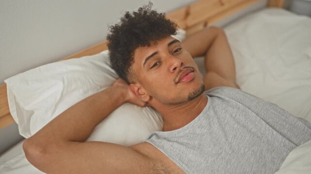 Man reclining on bed with arms behind head, smiling and showing teeth beside wooden headboard in a building; relaxed morning comfort.