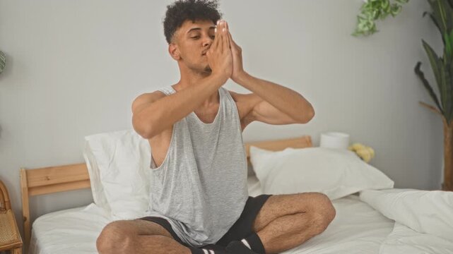 Man sitting crosslegged on bed with hands pressed in prayer gesture in studio during morning routine; calm mindfulness.
