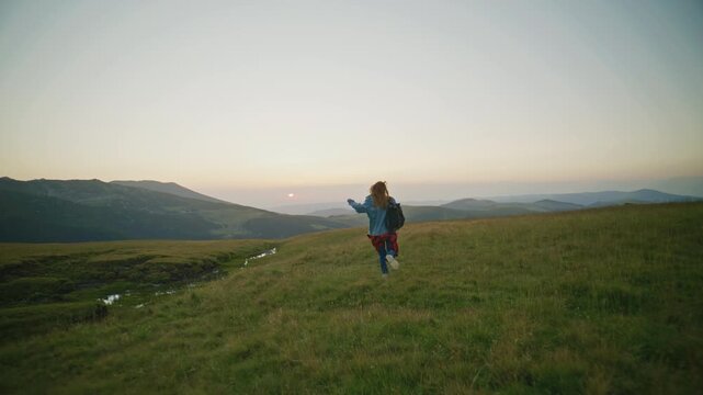 Person runs on a grassy hill at sunset with a backpack and mountain view in the background