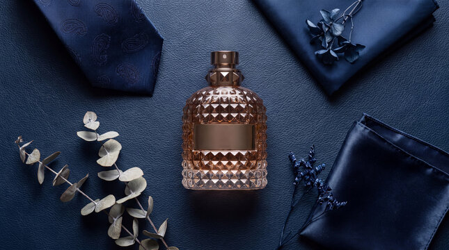 A high-end perfume bottle surrounded by a blue silk tie, pocket square, and dried floral accents on a dark navy blue textured leather surface.