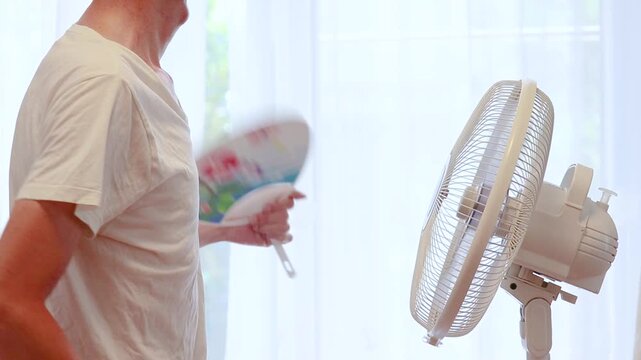 扇風機の風に当たりながらうちわを扇ぐ暑がりの男性
