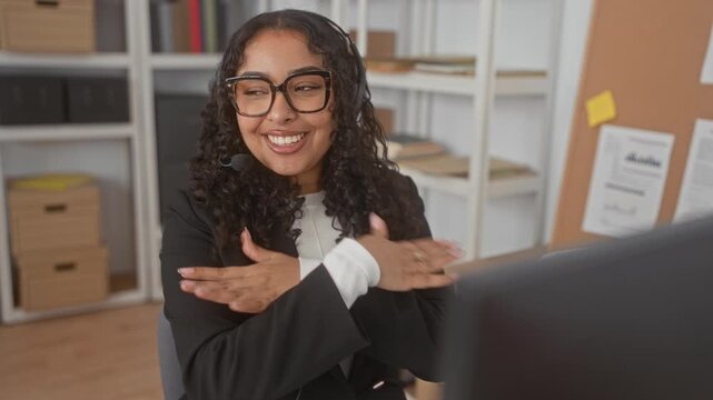 Woman wearing headset and glasses crosses arms hugging chest while smiling at computer monitor in building office desk area; joy gratitude.