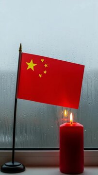 Chinese flag and red memorial candle on rainy window sill symbolizing mourning and remembrance