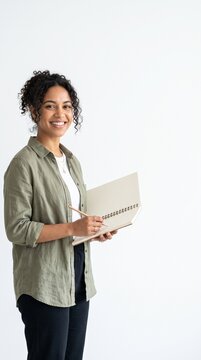 A smiling woman holding a notebook and a pencil, ready to work. Her hair is curly, and she is wearing a green shirt and black pants.
