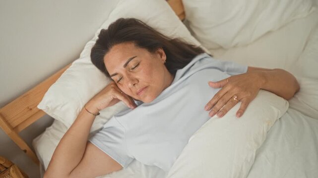 Woman middle age hispanic lying on bed with eyes closed, hand on pillow, fingers touching ring, soft expression in building; serene rest.