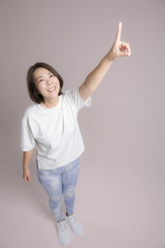 [ピンク背景]笑顔で指差す腕を高く伸ばして運動する女性、ダイエット成功や健康的で前向きな40代のミドル世代のフィットネスイメージ