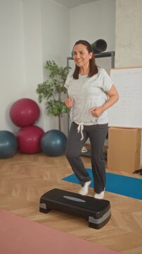 Woman stepping on an aerobic stepper with right foot raised in a light jogging motion, balancing near exercise balls and a whiteboard in a studio; wellness calm.