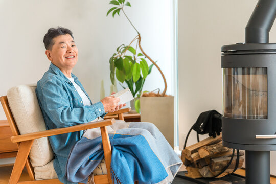 自宅のリビングの薪ストーブの前で椅子に座り本を読むシニア男性