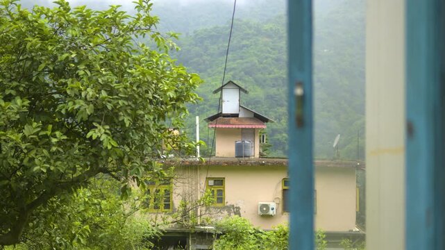A scenic establishing view framed by a window showing a traditional yellow house nestled among lush green slopes and foggy mountain mist in Mandi, Himachal Pradesh in the Indian Himalayas.